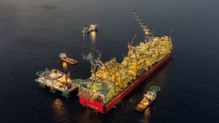 An offshore oil platform surrounded by support vessels floating in the sea. The platform is brightly lit and appears to be in operation, with smoke visible from its structures.