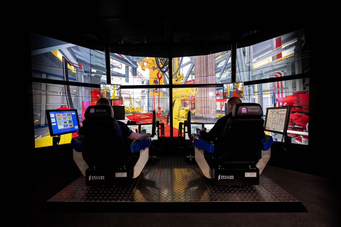 Two individuals are seated at a simulator displaying a virtual construction site with cranes and equipment on multiple screens. In front of them are control panels and a small touchscreen.