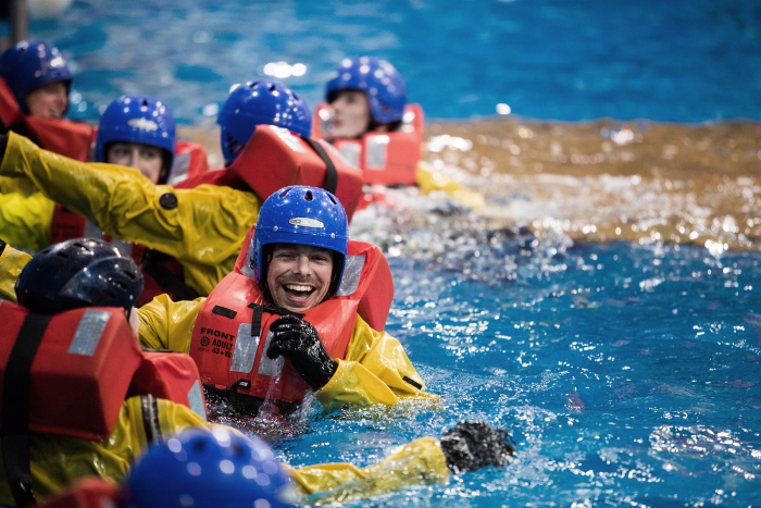 Individuals wearing life vests and helmets participate in water safety training, smiling and interacting in a pool.