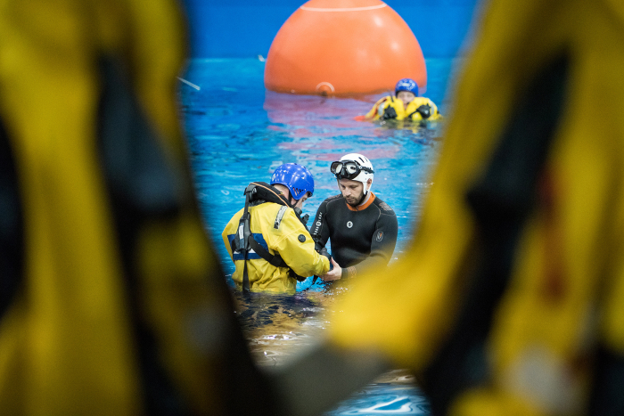 Two individuals in water activity gear are seen in the center of a pool, surrounded by others in yellow suits, with a large orange buoy in the background.