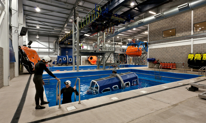 Indoor training facility with a large pool containing a helicopter simulator. People in wetsuits are practicing safety drills involving the submerged structure.