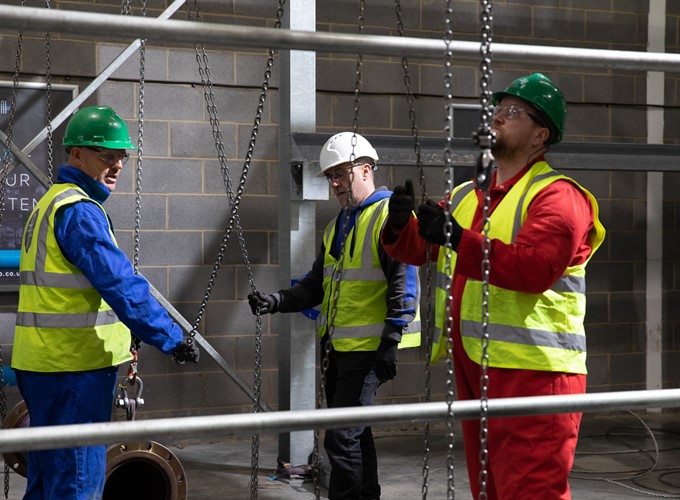 Three workers in high-visibility vests and hard hats operate chains and pulleys on a construction site.