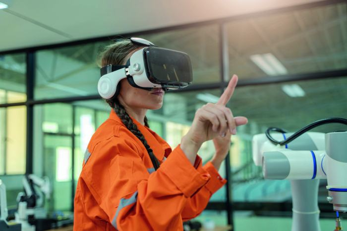 A person wearing an orange jacket and VR headset interacts with a robotic device in a modern industrial setting.