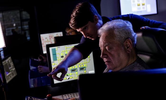 An older man operates controls while a younger man points at something on a screen in a control room setting with multiple monitors.