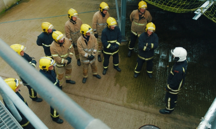 A group of firefighters dressed in yellow helmets and firefighting gear stands in formation, listening to another firefighter in white helmet giving instructions.