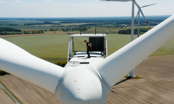 A technician is standing on top of a wind turbine in a rural area, performing maintenance. The surrounding landscape consists of expansive fields and distant tree lines.