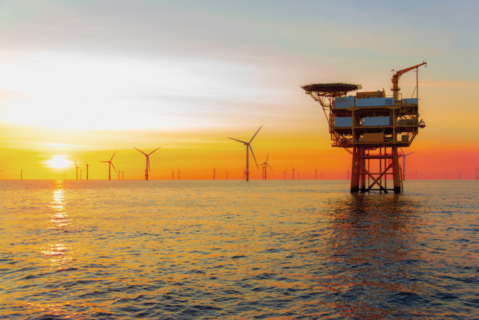 An offshore oil platform stands in the sea at sunset with wind turbines visible in the background.