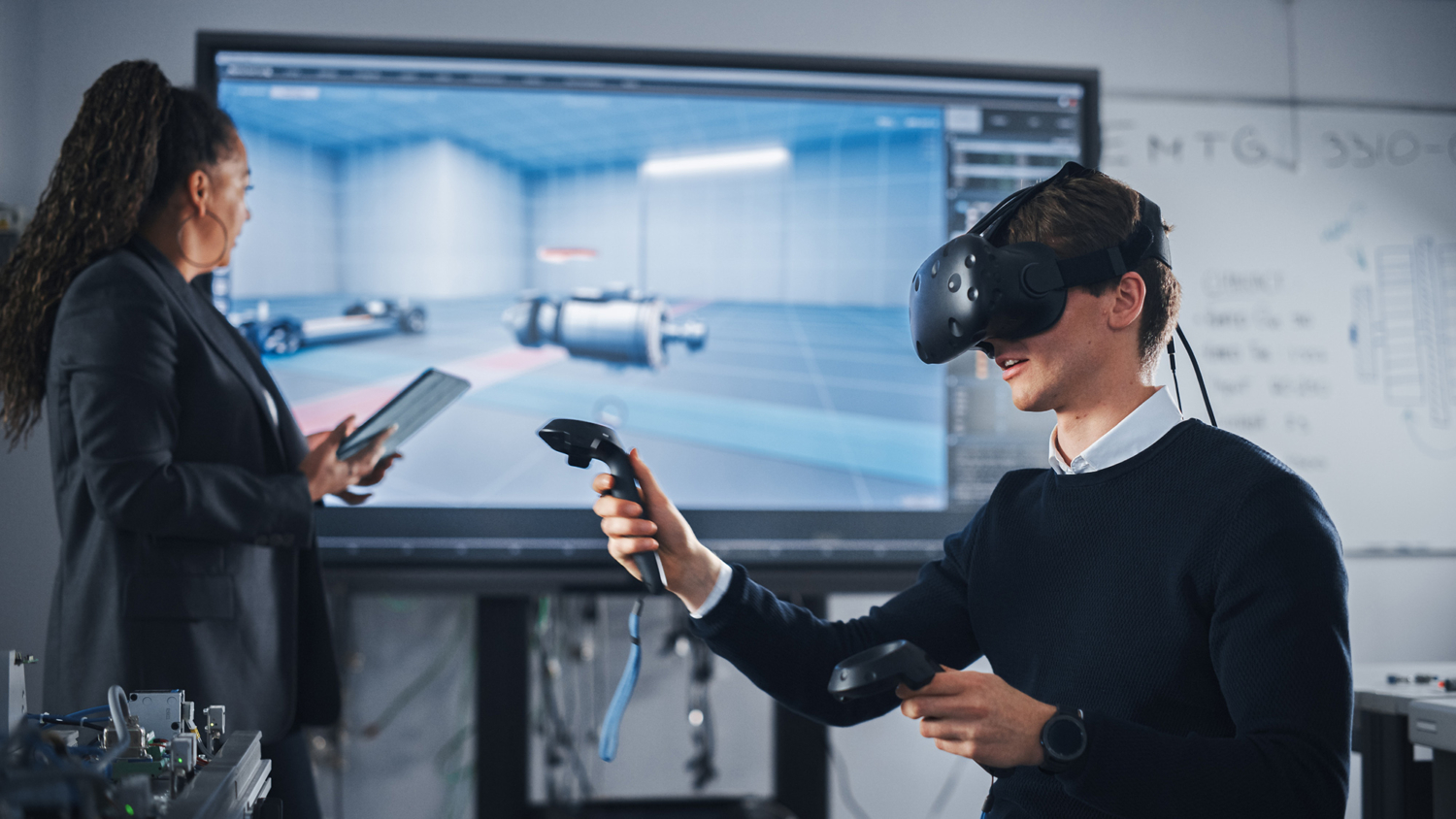 A person wearing a VR headset interacts with controllers while another person operates a tablet in front of a large screen displaying a 3D model in an office setting.