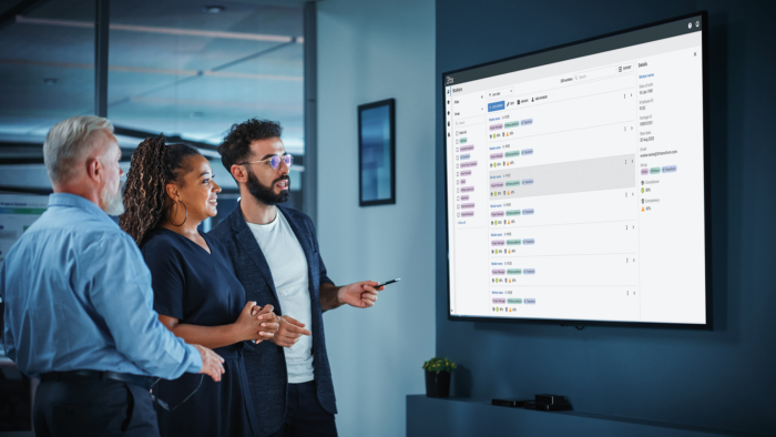 Three colleagues stand in front of a large screen displaying project management software, discussing the details and pointing at the screen.