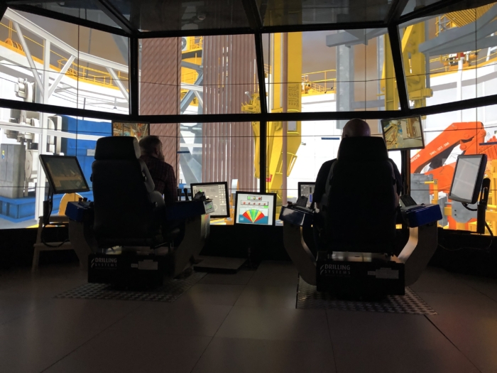 Two individuals operate computer systems in a control room with multiple monitors, overseeing heavy industrial machinery in the background.