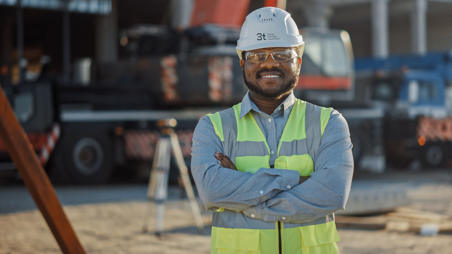 A construction worker in a safety vest and hard hat stands with arms crossed at a construction site.