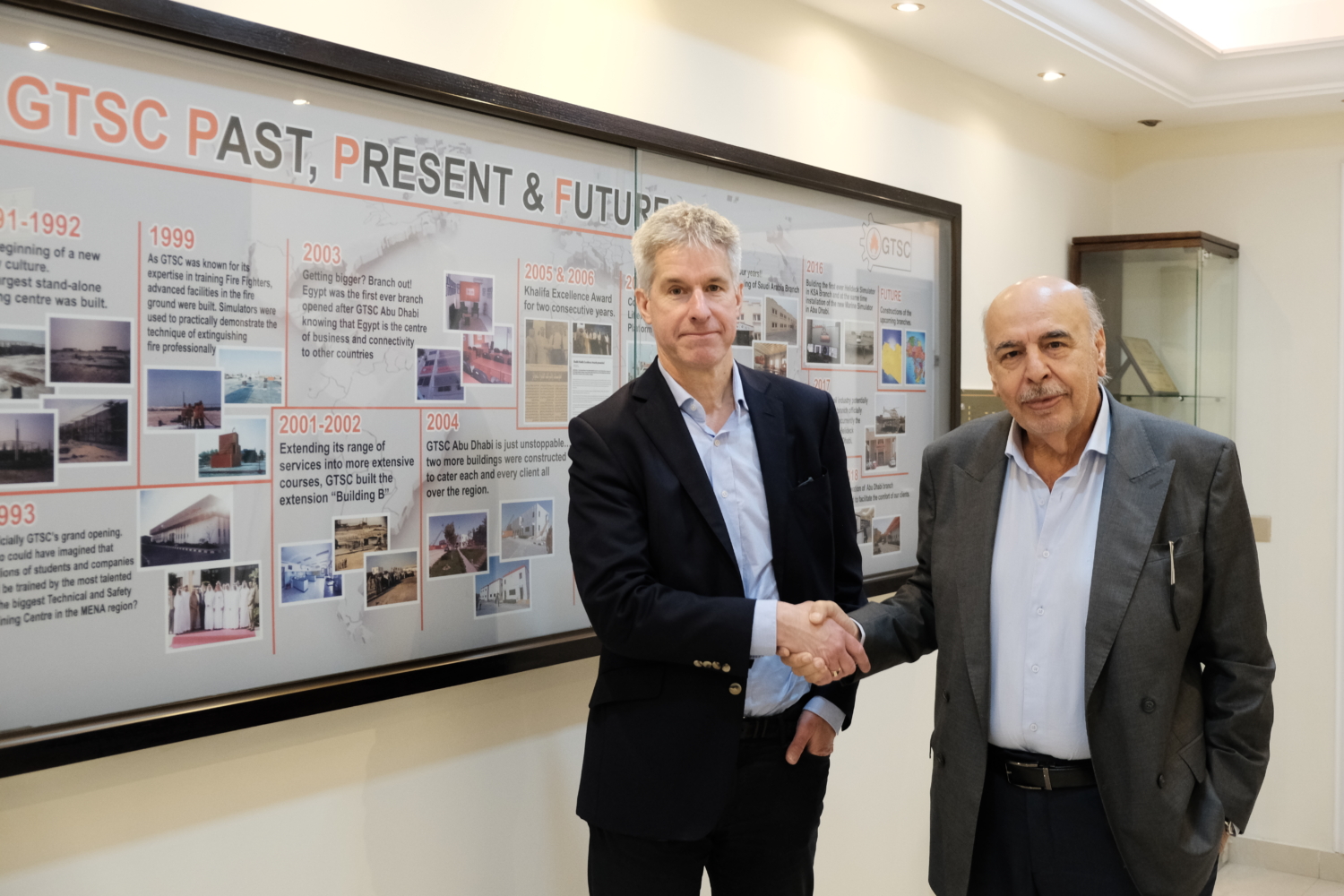 Two men shaking hands in front of a timeline display board labeled 