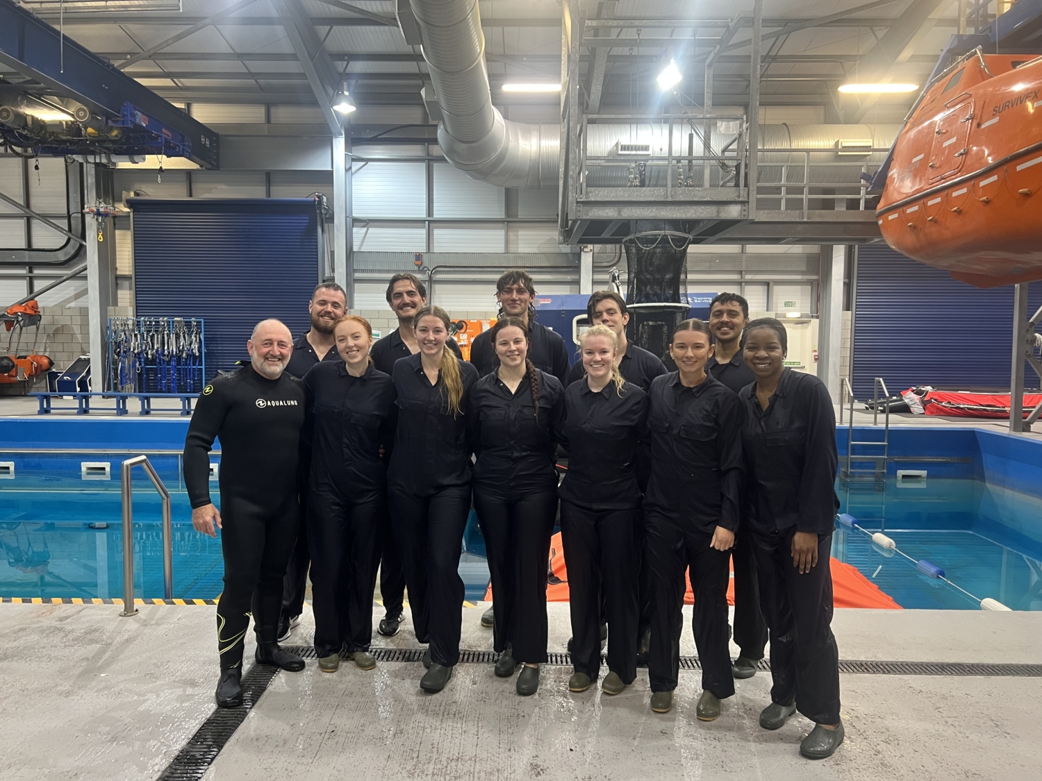 A group of people in black outfits stands together in an indoor facility near a pool and safety equipment, posing for a group photo.