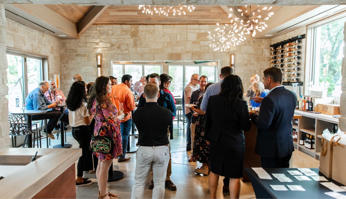 A group of people socializing in a spacious room with large windows, stone walls, and modern lighting, with wine bottles displayed on a rack in the background.